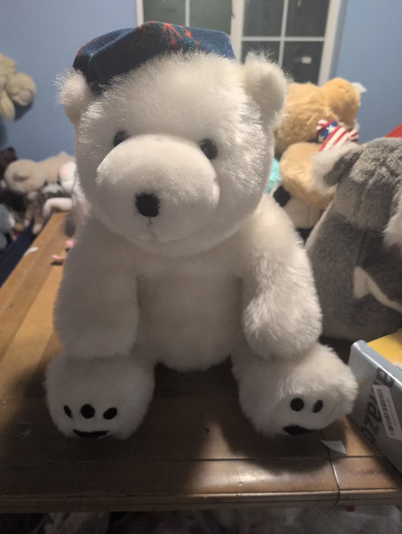 White plush polar bear sitting upright on a wooden table, wearing a navy and red tartan cap, with black-stitched paw pads and a black nose.