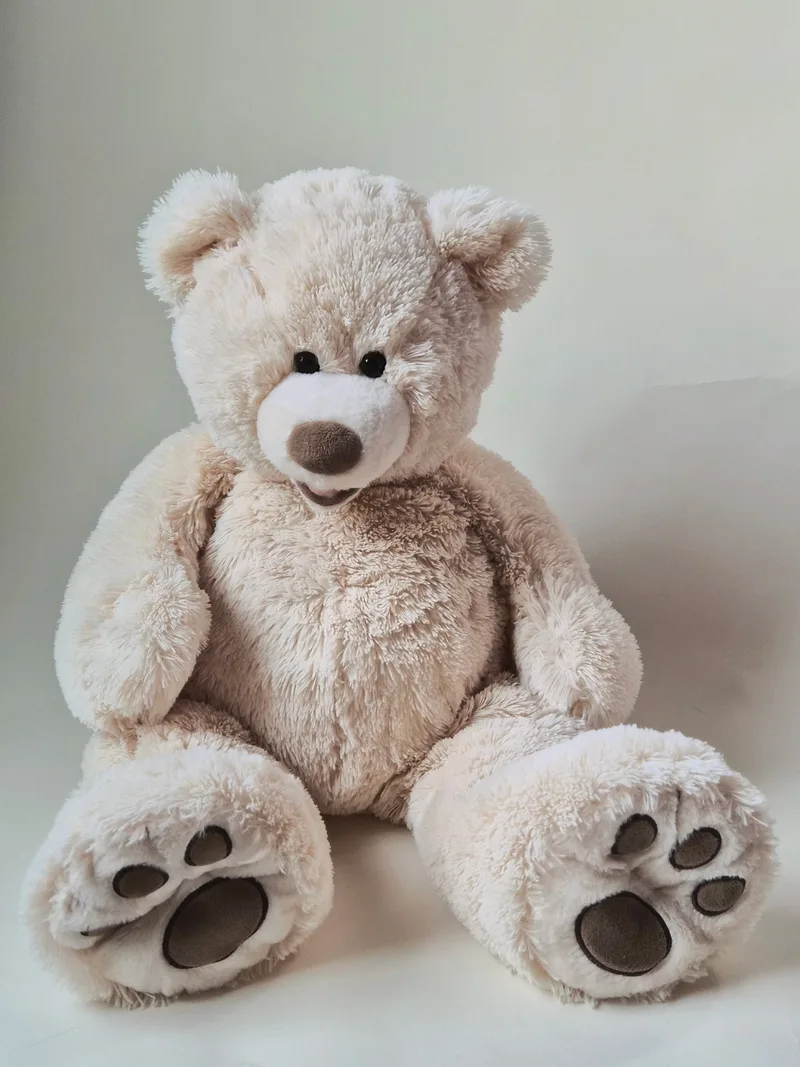 Large cream teddy bear with long shaggy fur sitting upright, showing black eyes, taupe nose, and oversized feet with embroidered brown paw pads