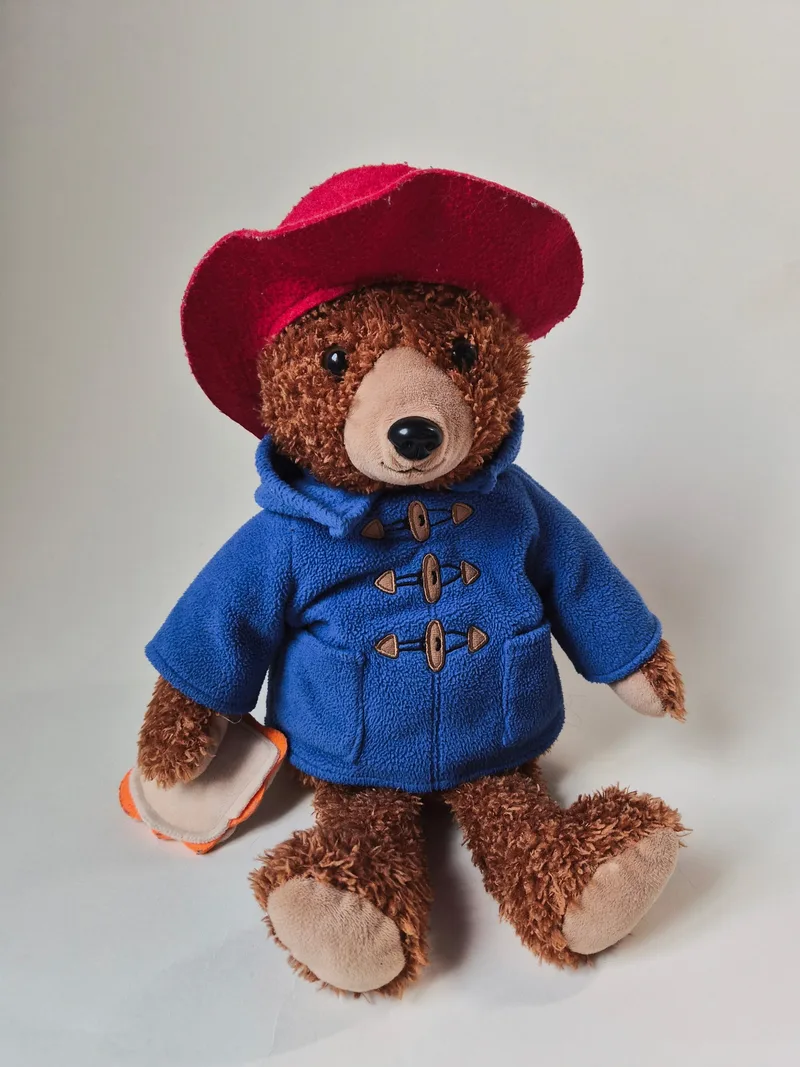 Plush Paddington bear with curly brown fur, wearing a red felt hat and blue toggle duffle coat, seated and holding an orange marmalade sandwich
