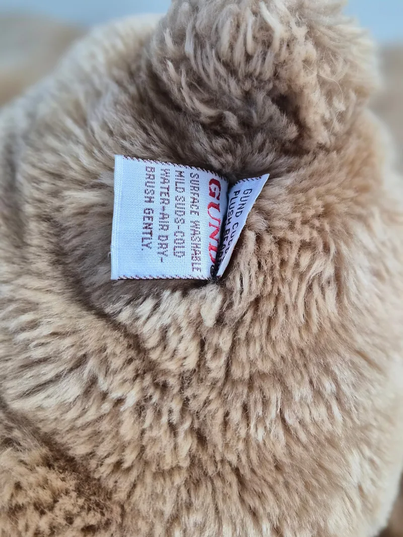 Close-up of a white Gund fabric tag with red lettering and care instructions, nestled in soft tan plush fur