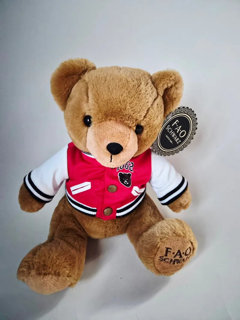 Caramel-brown FAO Schwarz teddy bear sitting upright, wearing a red and white varsity jacket with an 1862 patch, hang tags attached, FAO Schwarz embroidered on one paw pad