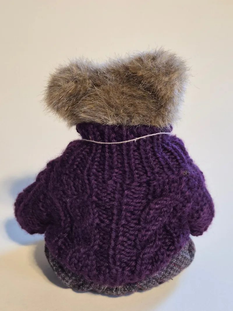 Back view of the teddy bear showing the plum cable-knit sweater and the top of gray tweed trousers
