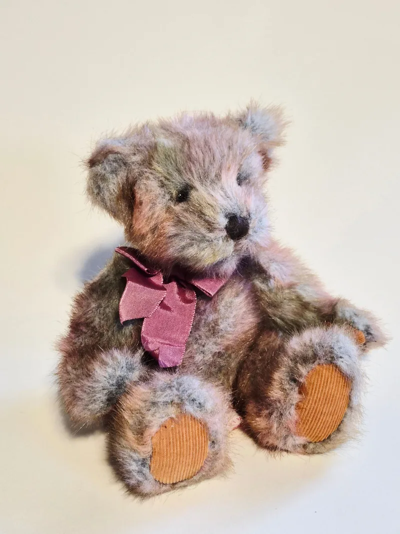 Small vintage teddy bear with multi-colored mauve, rose, and grey fur, a burgundy ribbon bow, and rust-orange corduroy paw pads, seated against a cream background