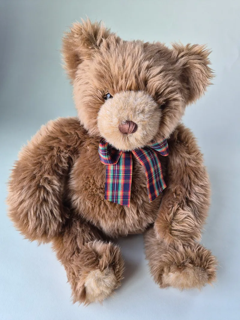 Front view of a shaggy honey-brown teddy bear wearing a tartan ribbon bow, seated with paws forward.