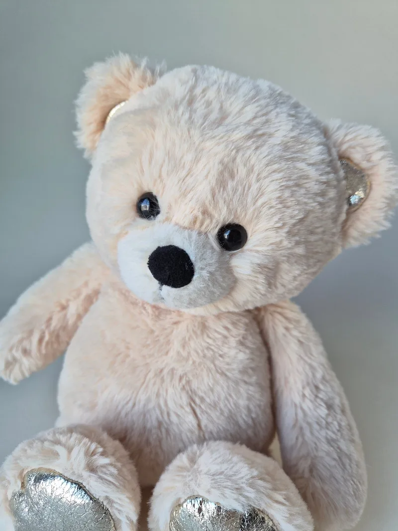 Close-up of a blush-cream teddy bear's face with black bead eyes, two-tone muzzle, and a silver ear tip