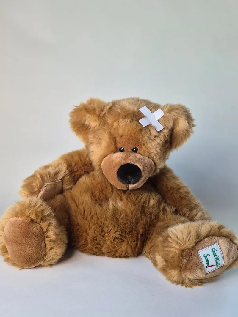Large honey-brown teddy bear sitting upright with a white felt bandage taped to his forehead and an embroidered patch on one foot
