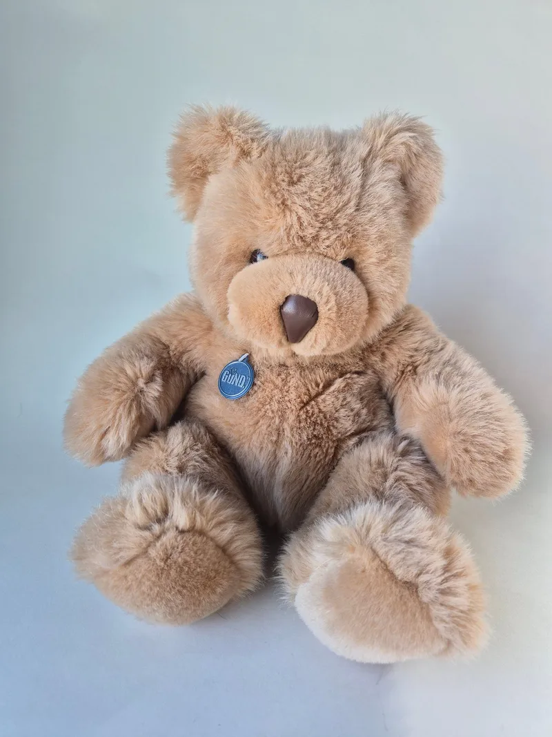 Honey-caramel Gund teddy bear seated, with dark eyes, suede-brown nose, and a blue oval Gund charm at the chest