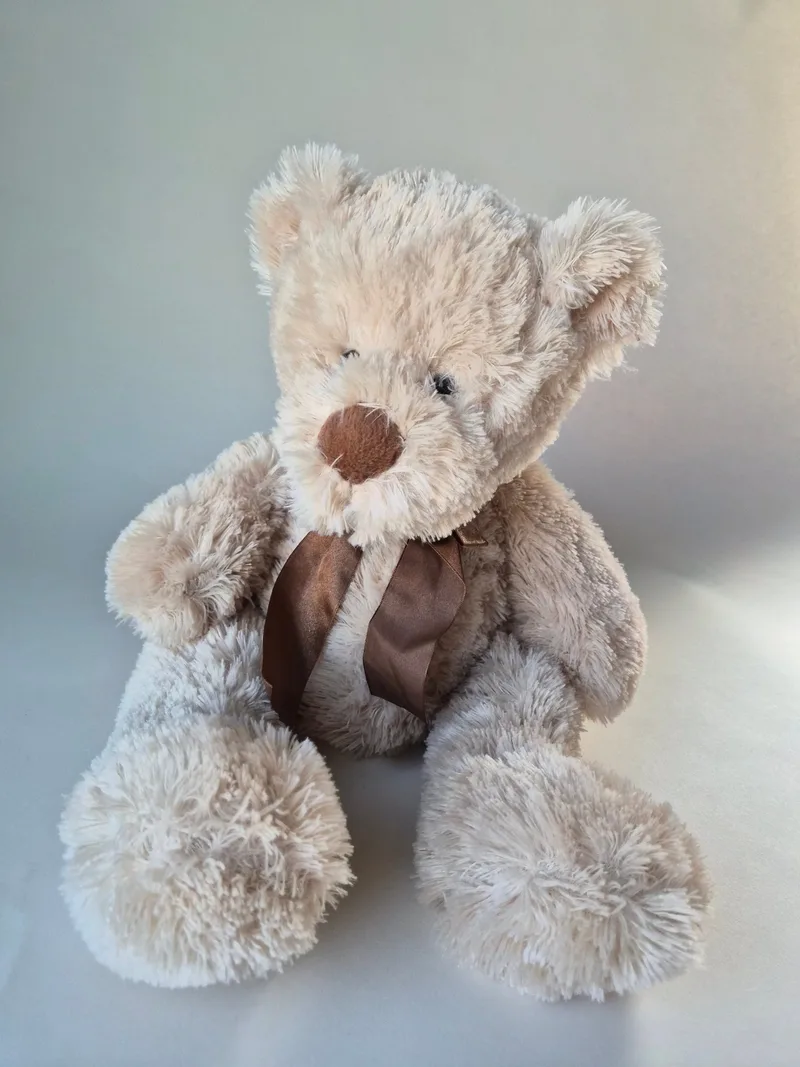 Full seated view of an oat-and-cream shaggy teddy bear with large paw pads and a brown satin neck ribbon