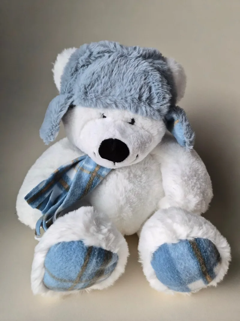 Seated white polar bear plush in a grey-blue faux-fur trapper hat with earflaps, blue plaid fleece scarf, and plaid fleece pads on both feet