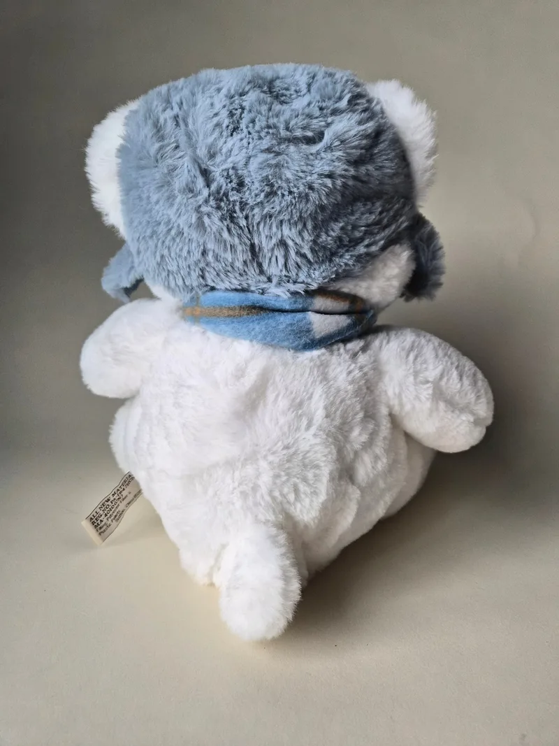 Side and back view of the white polar bear plush showing the fitted trapper hat, scarf knot, and a fabric tag at the lower back seam