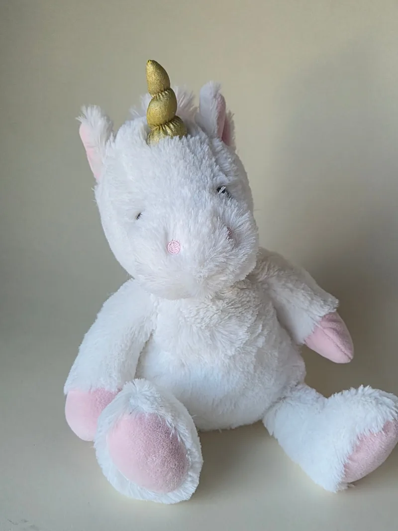 Front view of a white plush unicorn seated, with a gold twisted horn, pink-lined ears, embroidered pink nose and rose-colored paw and heel pads.