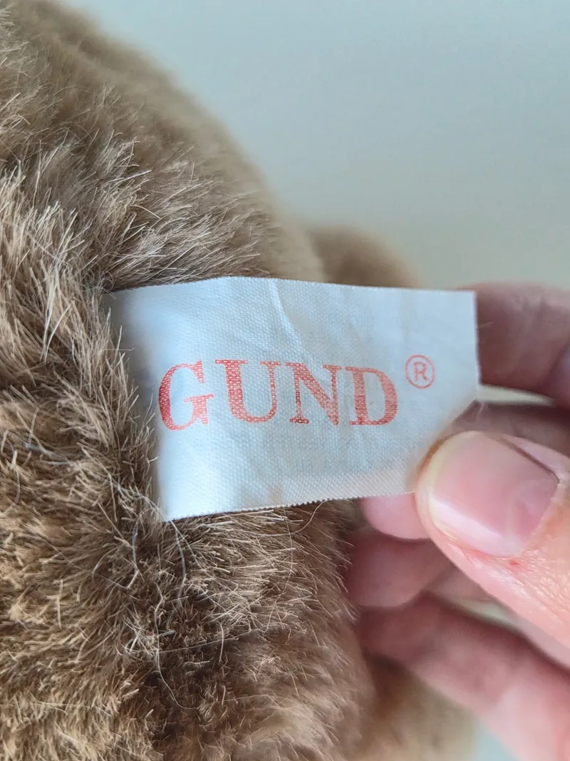 A hand holding the bear's white woven label printed with GUND in red script