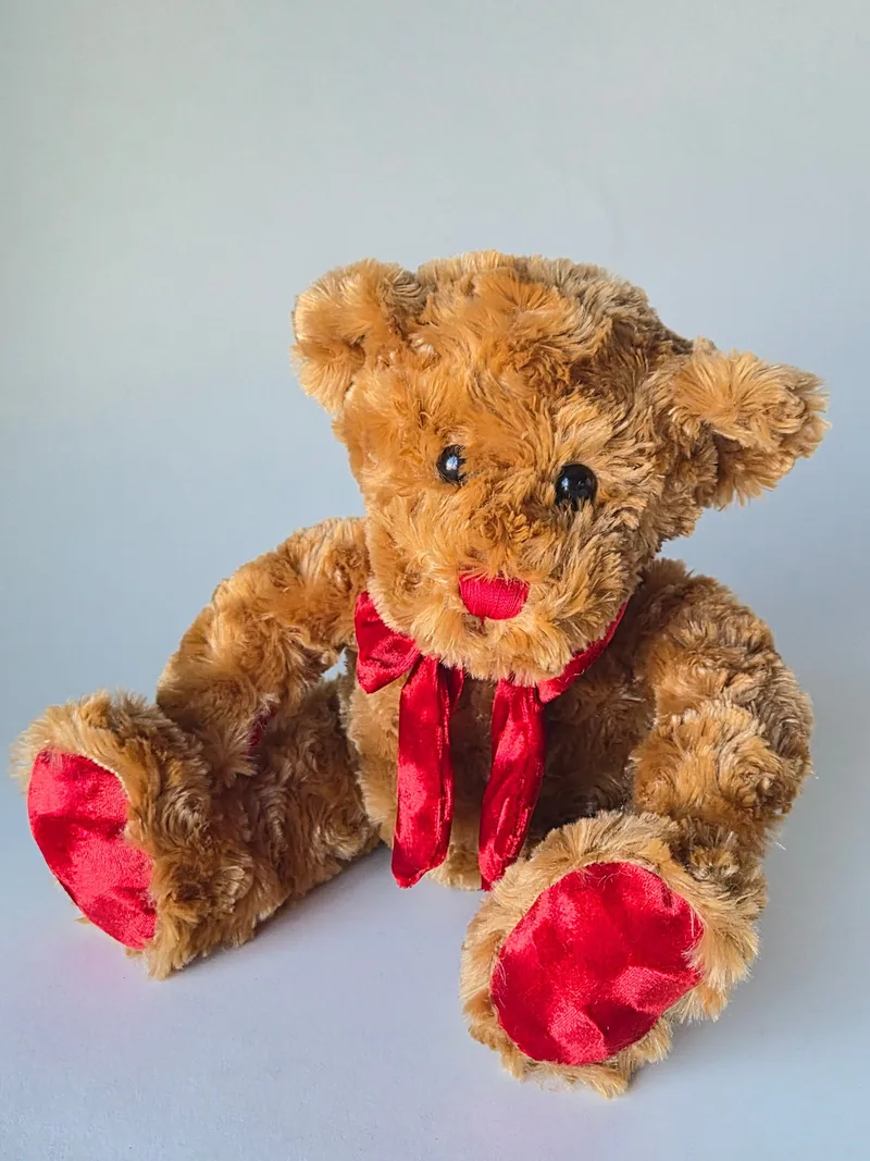 Curly honey-brown teddy bear sitting with legs out, showing red satin foot pads and a red satin bow at the neck