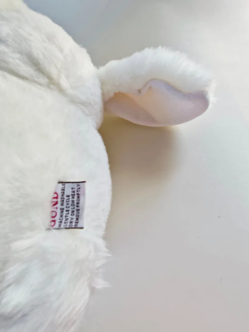 Detail of one ear turned to show the pale pink lining with a faint lilac mark, and the Gund care label reading machine washable, gentle cycle, dry on low heat, remove promptly