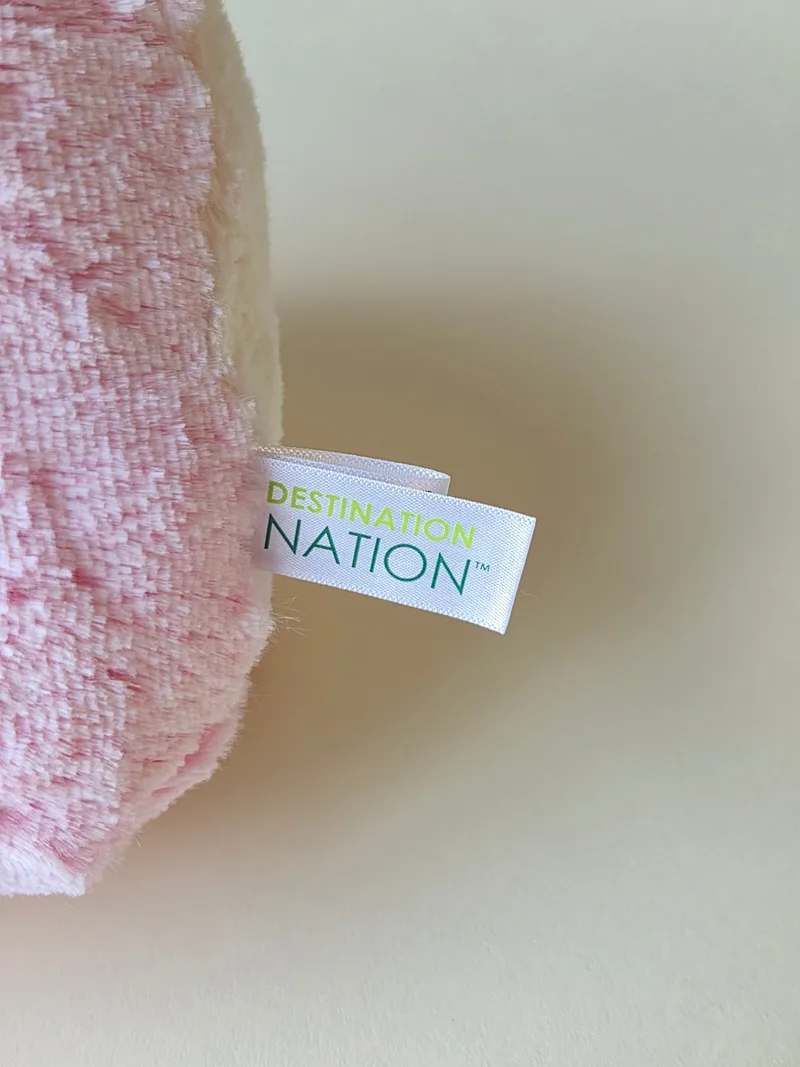 Close-up of a white satin label sewn into the pink plush, reading DESTINATION NATION in green lettering