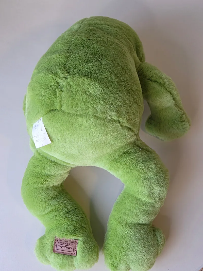 Back view of the green frog lying face down, showing the printed white care label at the back seam and darker pressed plush across the shoulders