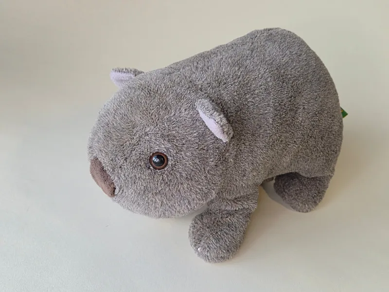 Grey plush wombat lying low, three-quarter view, with suede ears and amber eyes