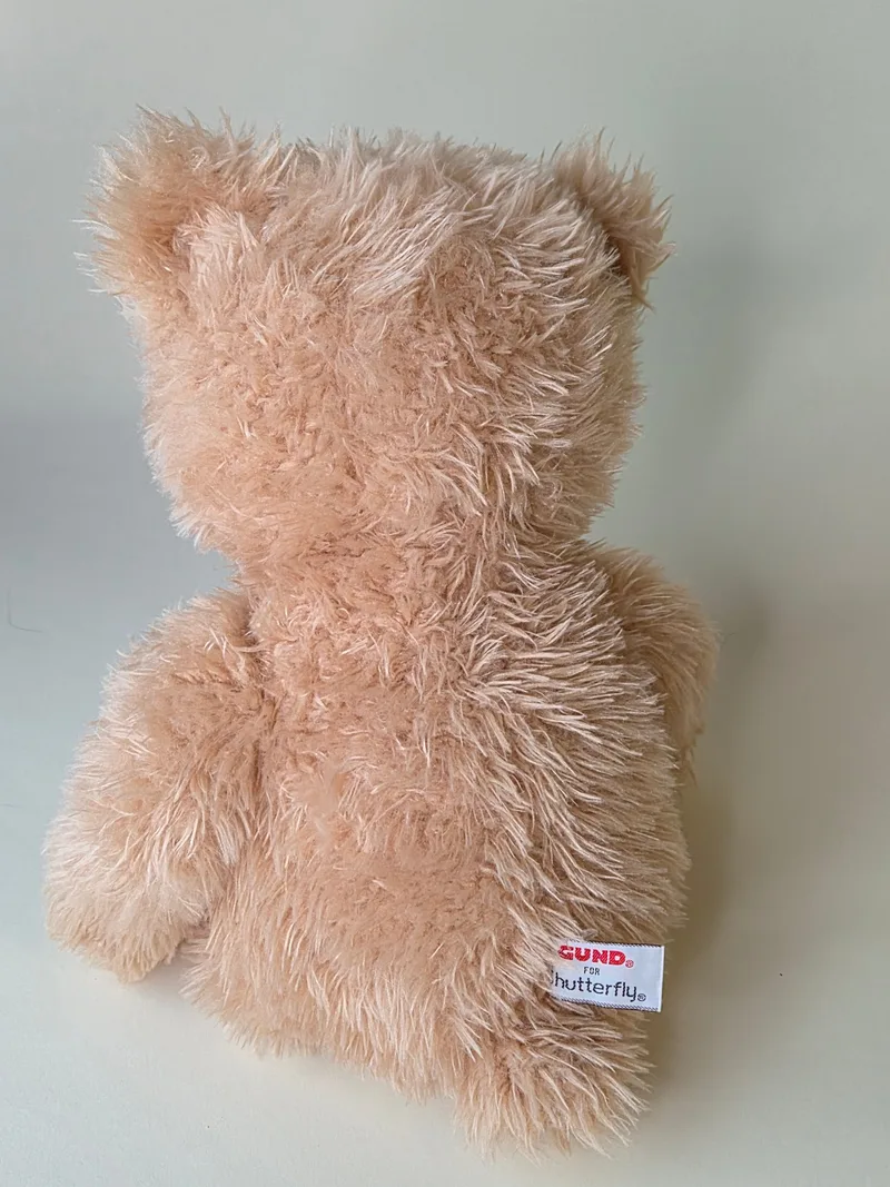 Side-back view of the apricot teddy bear showing long pale pile and a white maker's tag in the lower seam
