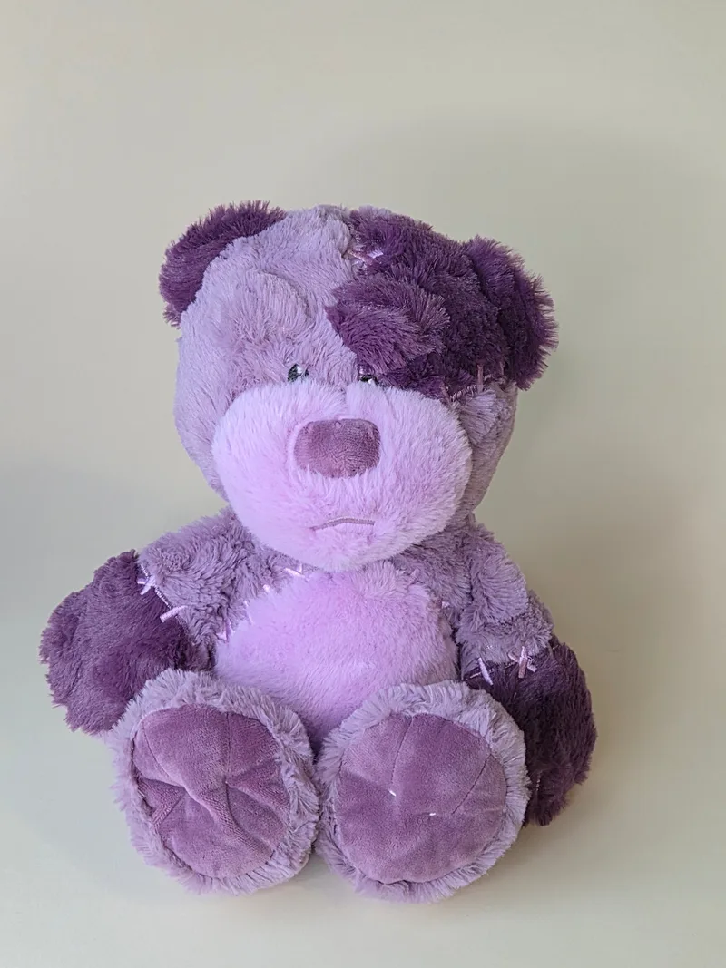 Front view of a lilac and aubergine two-tone teddy bear with lavender X-stitch patches and a stitched straight mouth