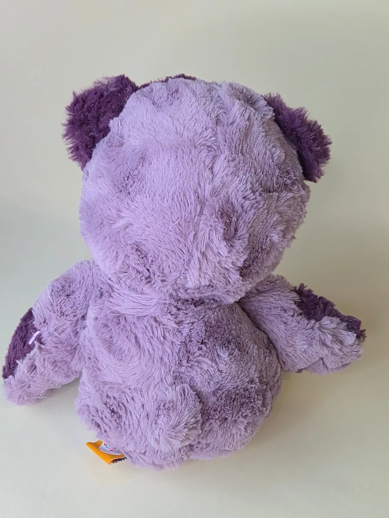 Back view of the purple teddy bear showing dense plush and an orange woven hem tag at the lower back