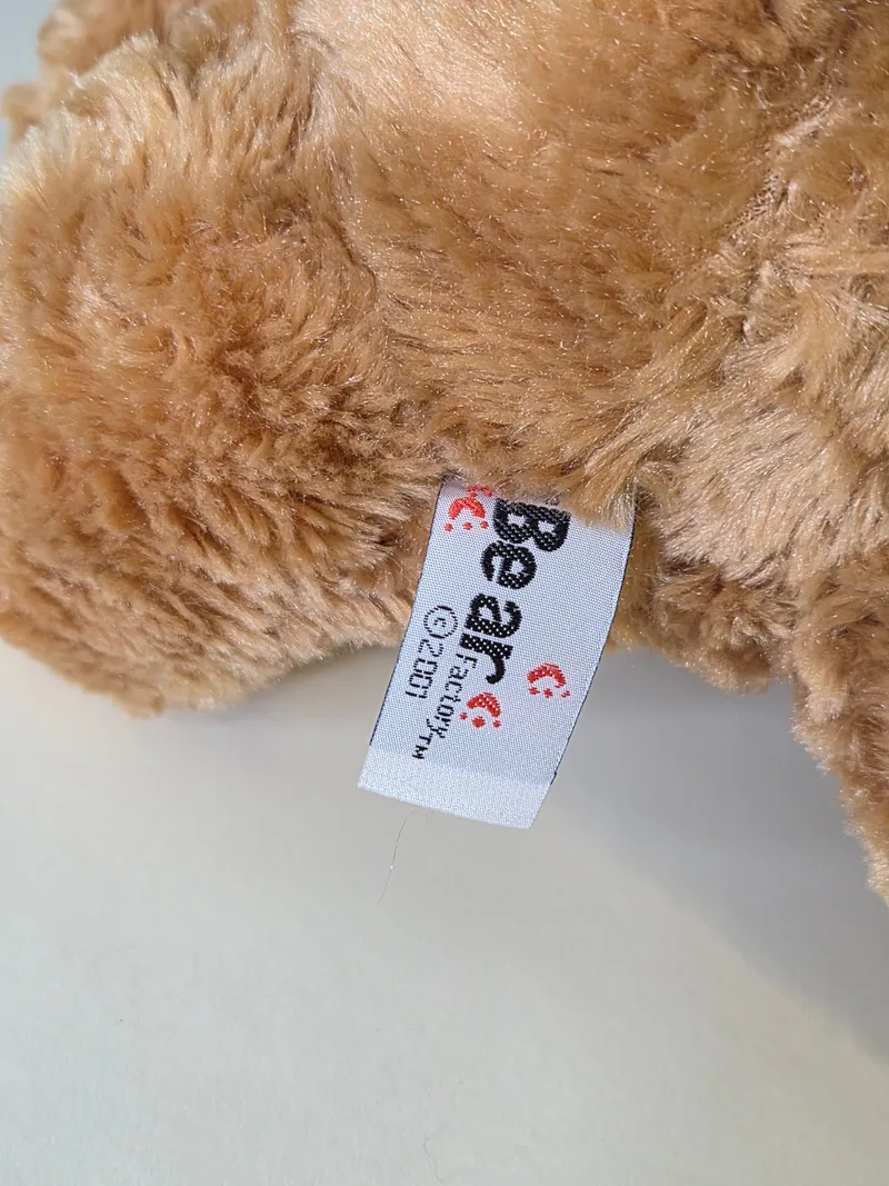 Close-up of the woven hem tag reading 'Bear Factory © 2001' tucked into the bear's fur.