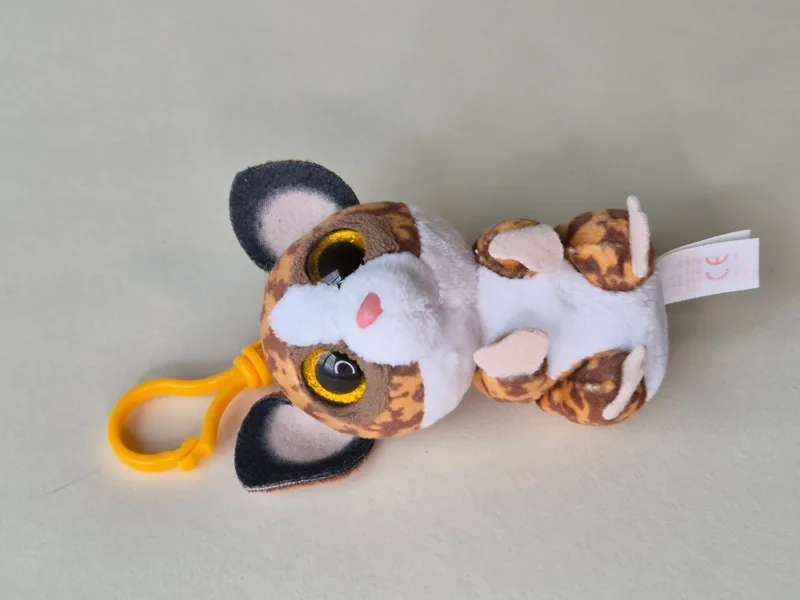 Same keychain plush lying on its back, showing the yellow clip, pale paw pads, and a fabric tag sewn into the side seam.