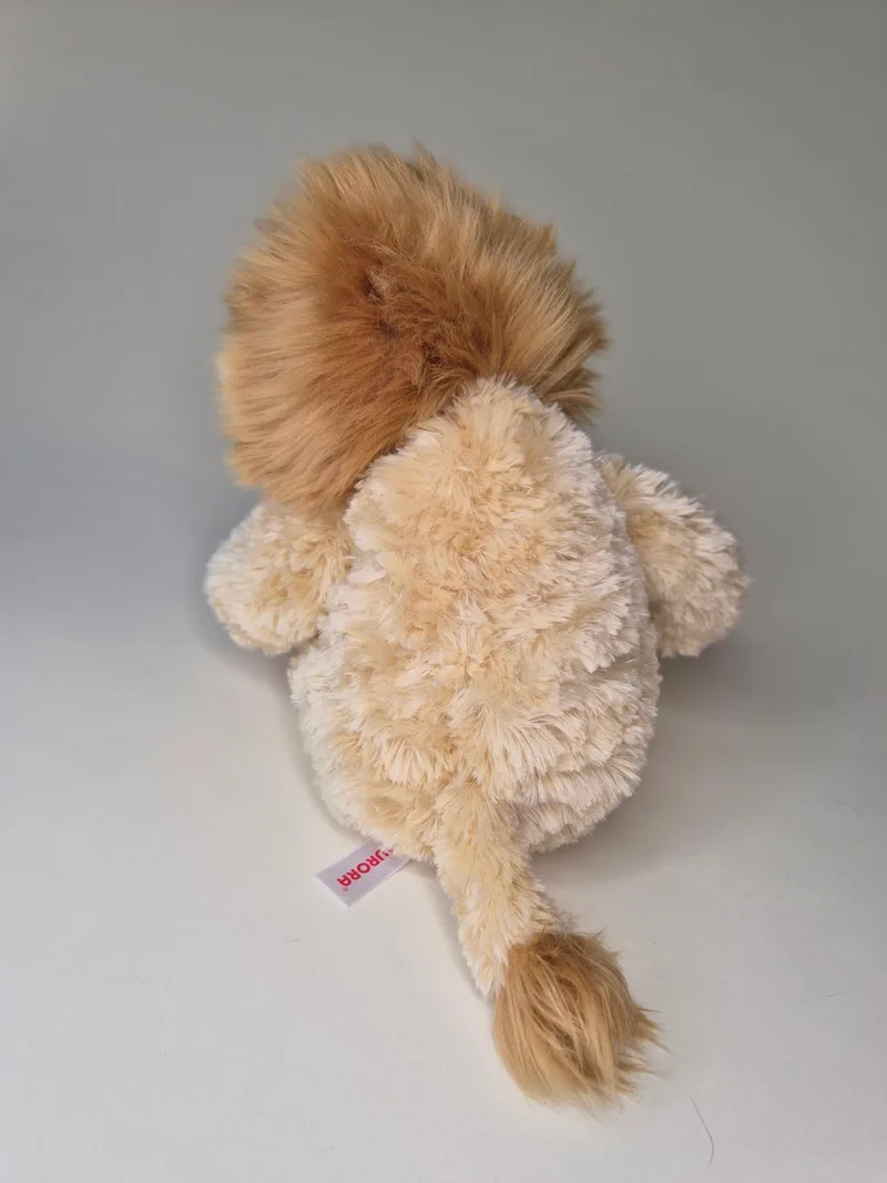 Back view of the same plush lion showing the full spread of the mane, a small tufted tail, and a woven Jurora fabric tag stitched into the lower back seam.