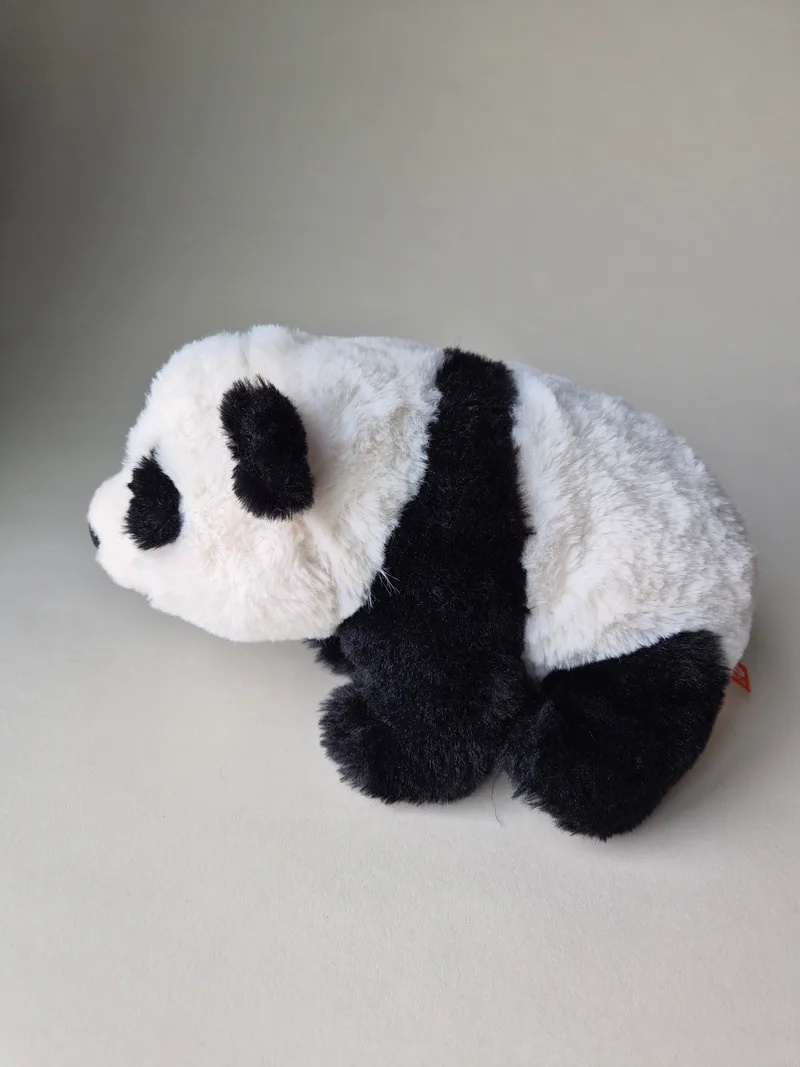Side view of a crouched black-and-white panda plush with charcoal eye patches against a grey background
