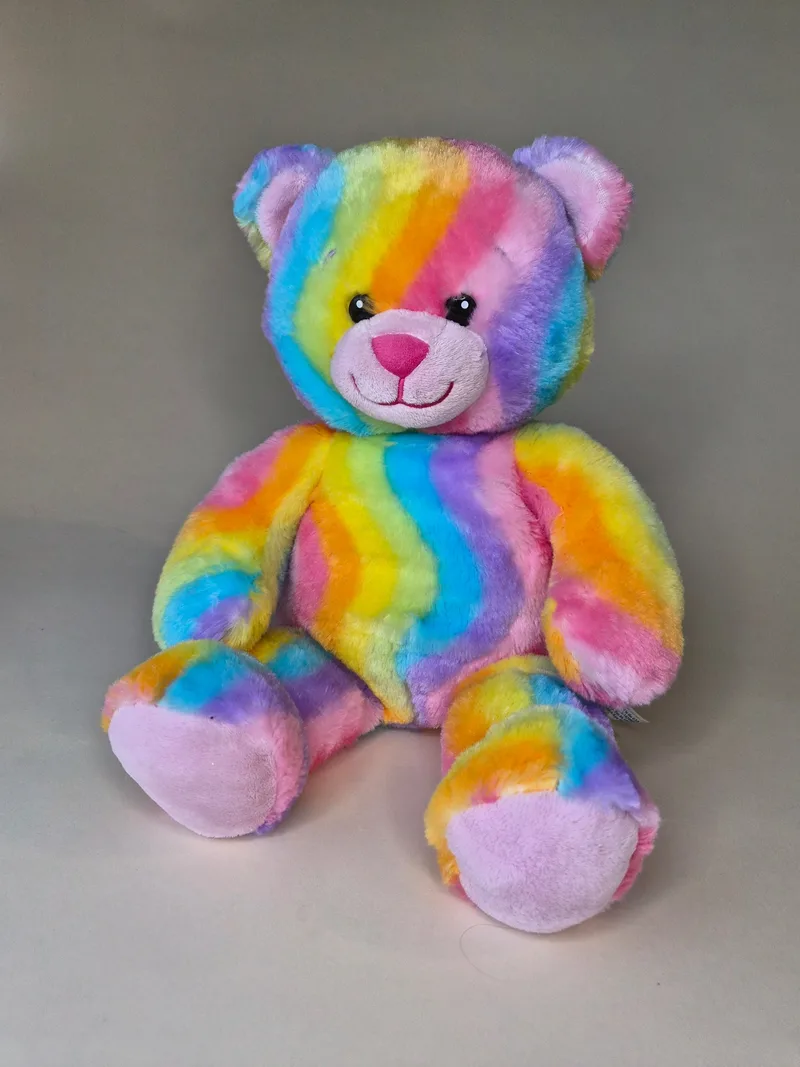 Front view of a rainbow tie-dye teddy bear with pink nose, ears, and paw pads, seated against a gray backdrop