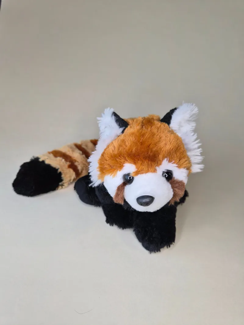 Front three-quarter view of a plush red panda lying down, showing white-rimmed eyes, black nose, and rust-orange and cream fur