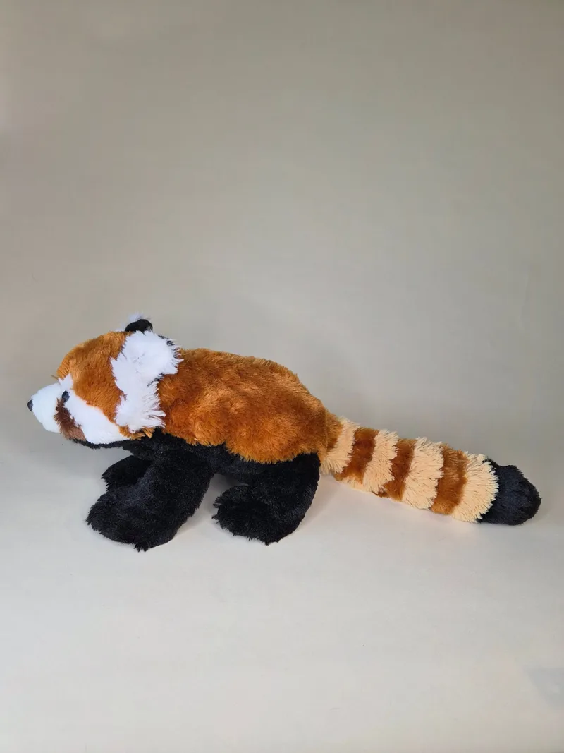 Side profile of the red panda plush lying with legs tucked beneath its body, tail extended alongside